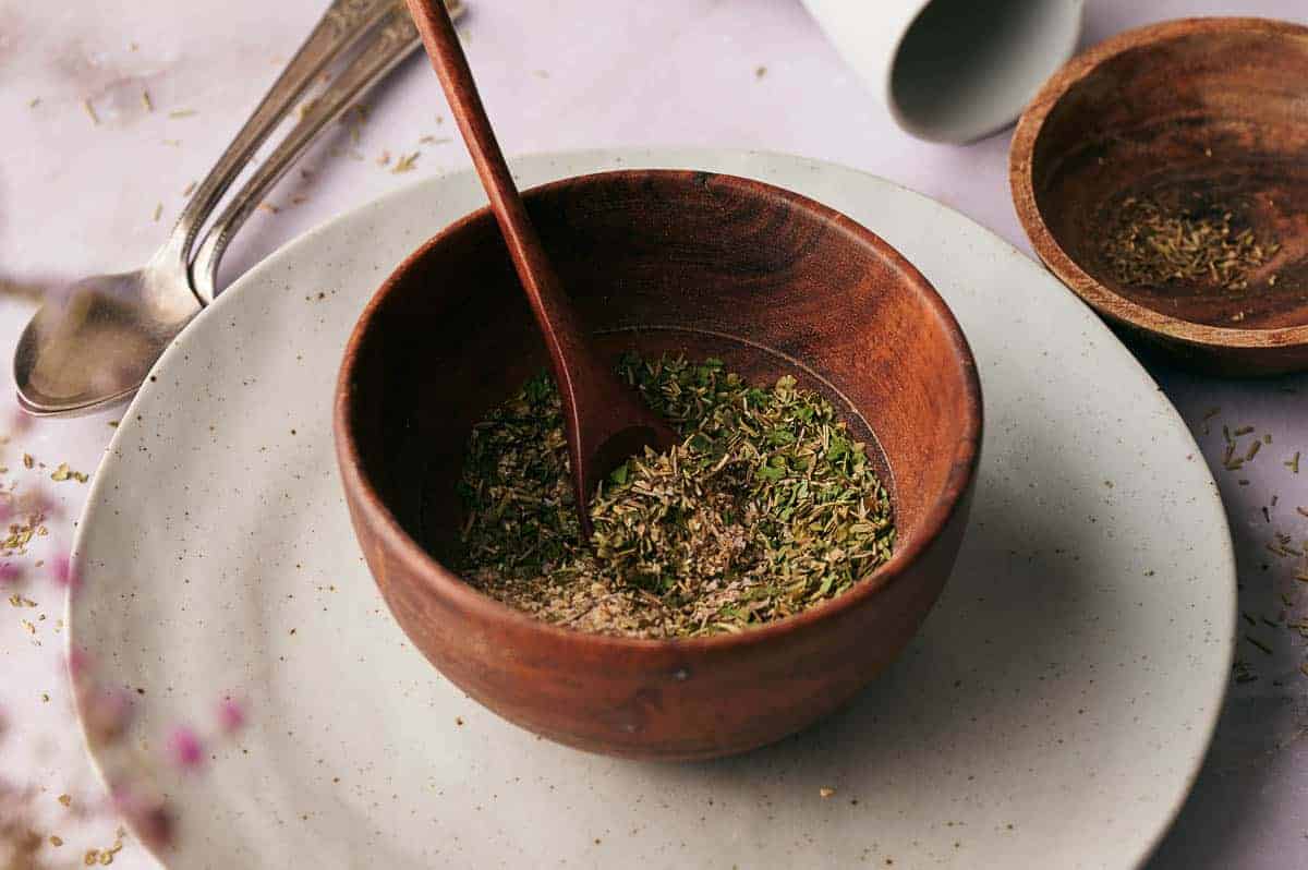 A wooden bowl filled with pizza seasoning on a plate.