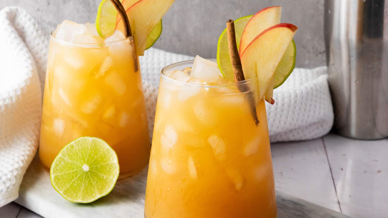 Two glasses of apple cider mocktail garnished with apple slices, lime slices, and cinnamon sticks.