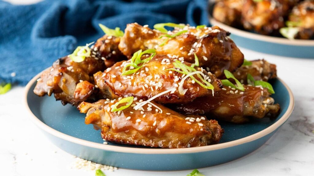A plate of glazed chicken wings garnished with sesame seeds and sliced green onions, set on a blue plate with a blue cloth in the background.