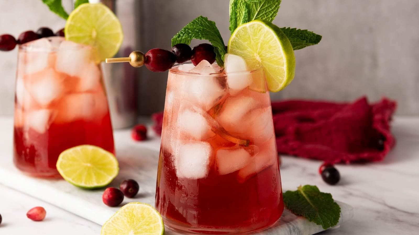 Two glasses of red iced drinks garnished with lime slices, mint leaves, and skewered cranberries, set on a marble surface.