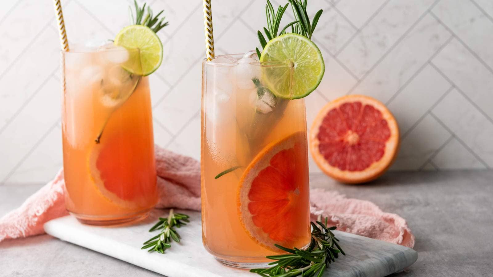 Two tall glasses of iced grapefruit mocktail garnished with lime slices, rosemary sprigs, and grapefruit slices, set on a marble tray with a halved grapefruit in the background.