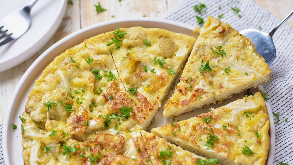 A sliced potato frittata garnished with chopped parsley on a white plate, with one slice being served.