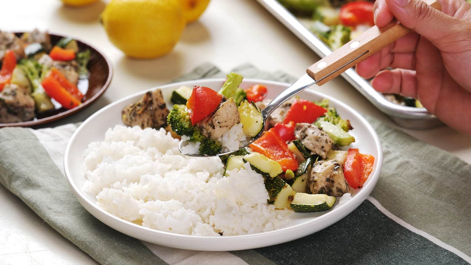 A hand serves roasted vegetables and chicken onto a plate of white rice; lemons and more food are visible in the background.