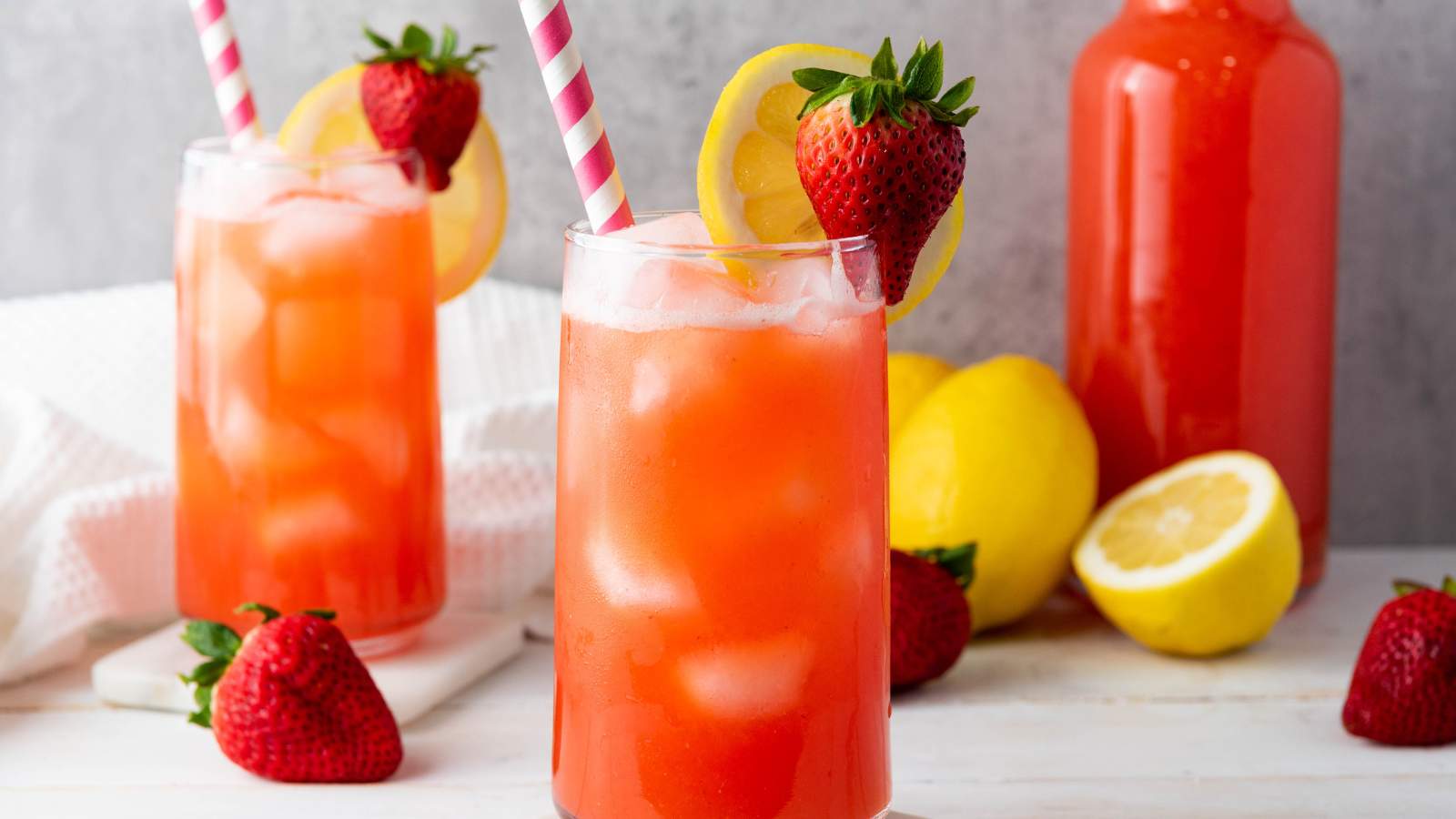 Two glasses of strawberry lemonade with ice, lemon slices, strawberries, and striped straws.