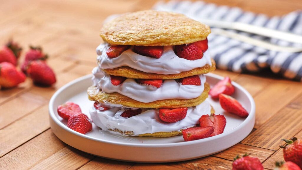 A stack of three pancakes layered with whipped cream and sliced strawberries, served on a white plate with extra strawberries on the side.
