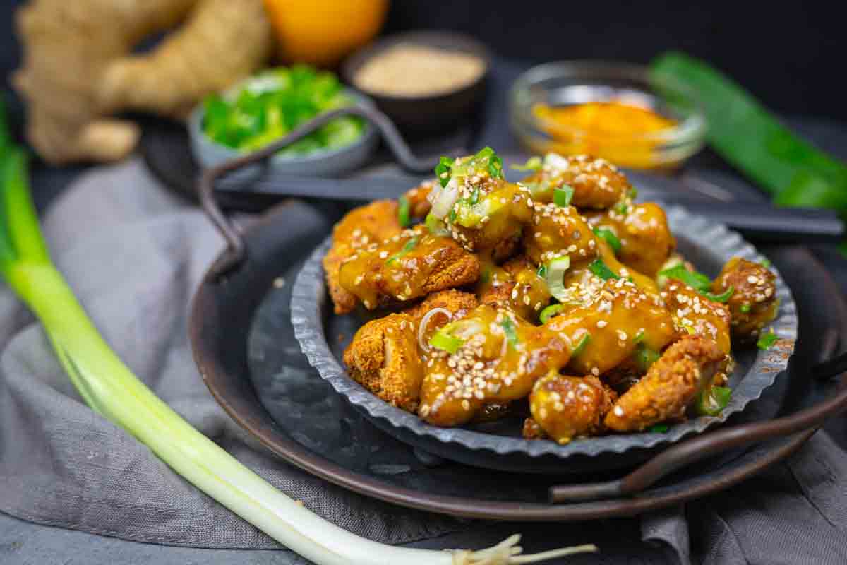 A plate of sesame-coated, sauced chicken pieces garnished with sliced green onions. Various fresh ingredients and spices are visible in the background.