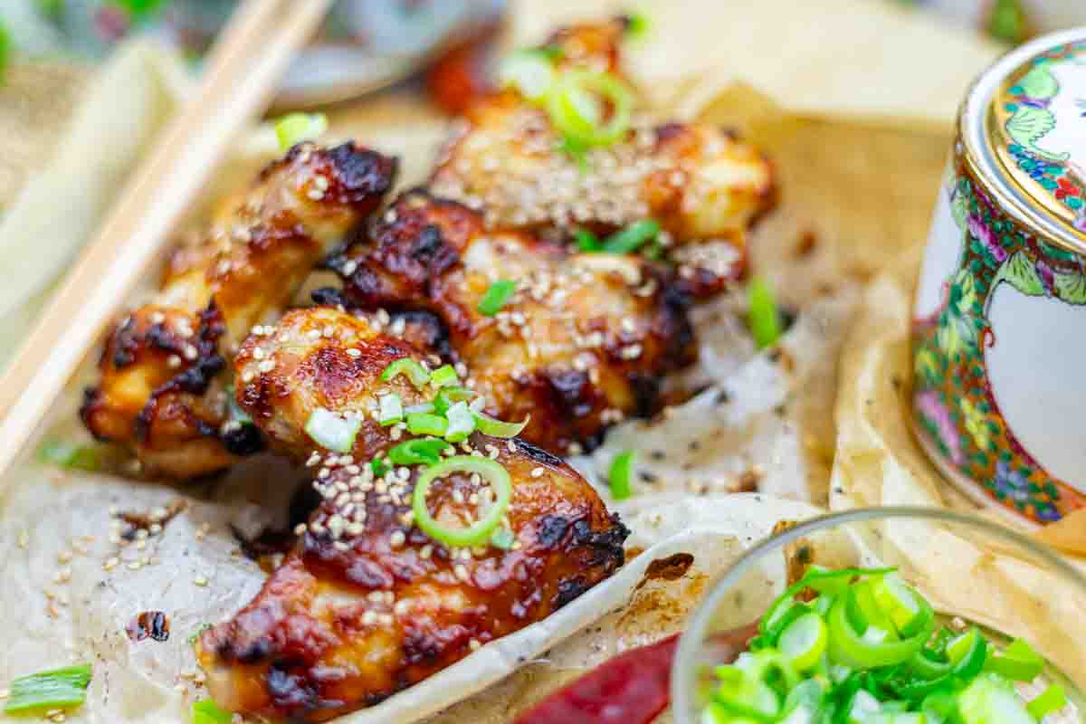 BBQ chicken wings topped with sesame seeds and chopped green onions, served on parchment paper with a side dish partially visible.