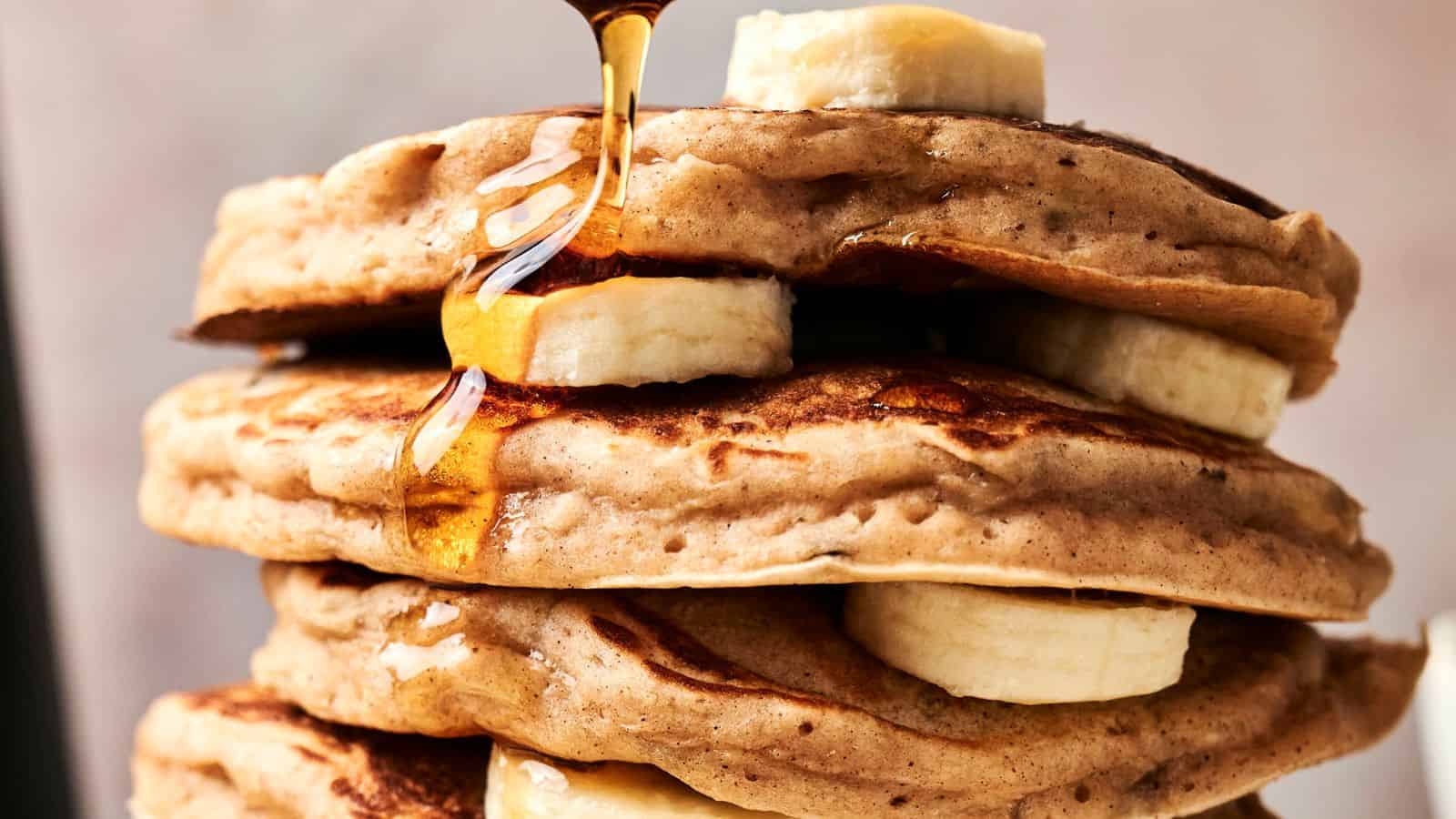 A stack of pancakes with banana slices between layers, topped with more banana and being drizzled with syrup.