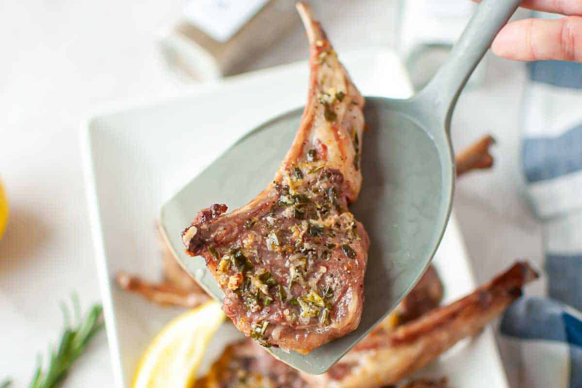 A cooked lamb chop seasoned with herbs is lifted on a gray spatula above a white plate with more lamb chops and lemon slices.