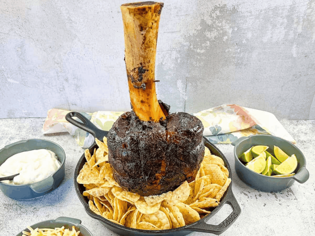 A large roasted meat shank on a bone, displayed upright in a skillet surrounded by nachos, with bowls of lime wedges and sauce nearby.