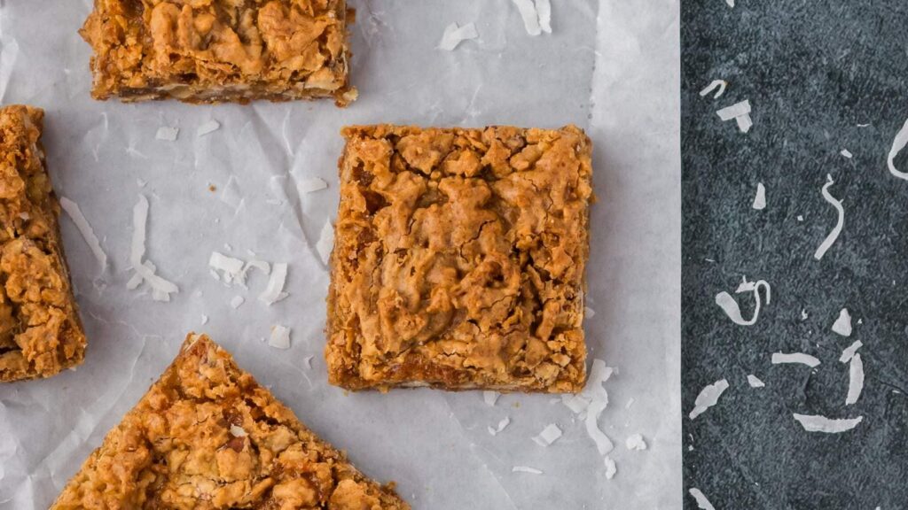 Three butterscotch brownies on parchment paper with shredded coconut scattered nearby on a dark surface.