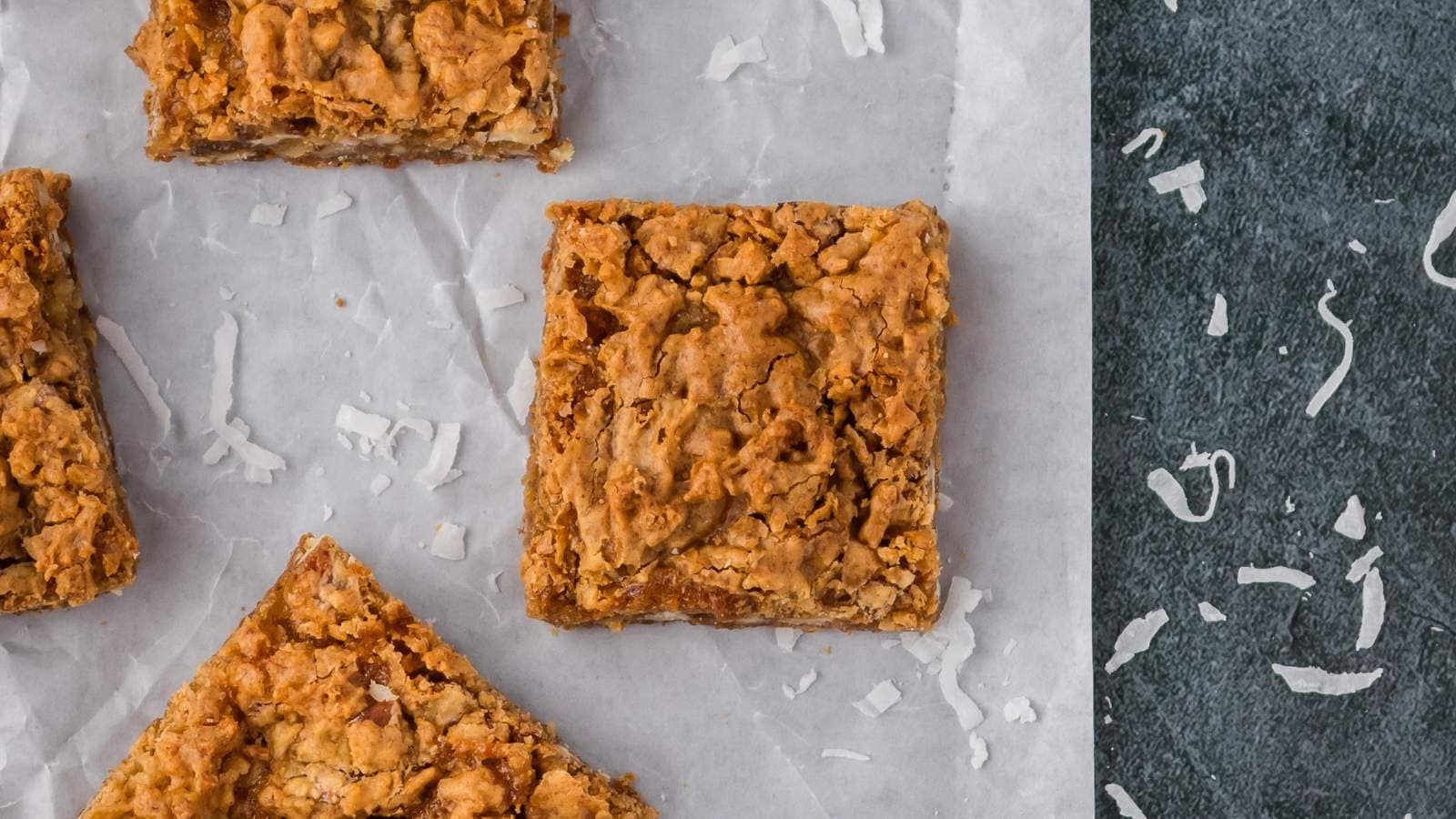 Three butterscotch brownies on parchment paper with shredded coconut scattered nearby on a dark surface.