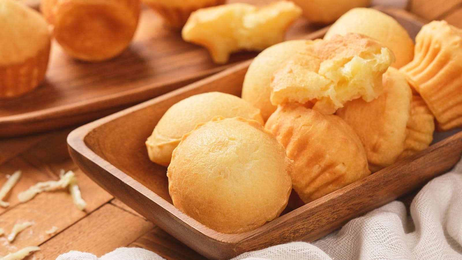A wooden tray filled with round, golden cassava breads and muffins, some with bites taken out, placed on a wooden surface beside a white cloth.