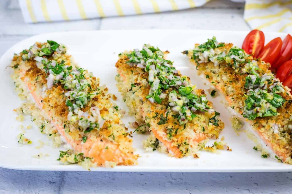 Three pieces of breadcrumb-crusted baked salmon topped with chopped herbs and served on a white plate with sliced cherry tomatoes.