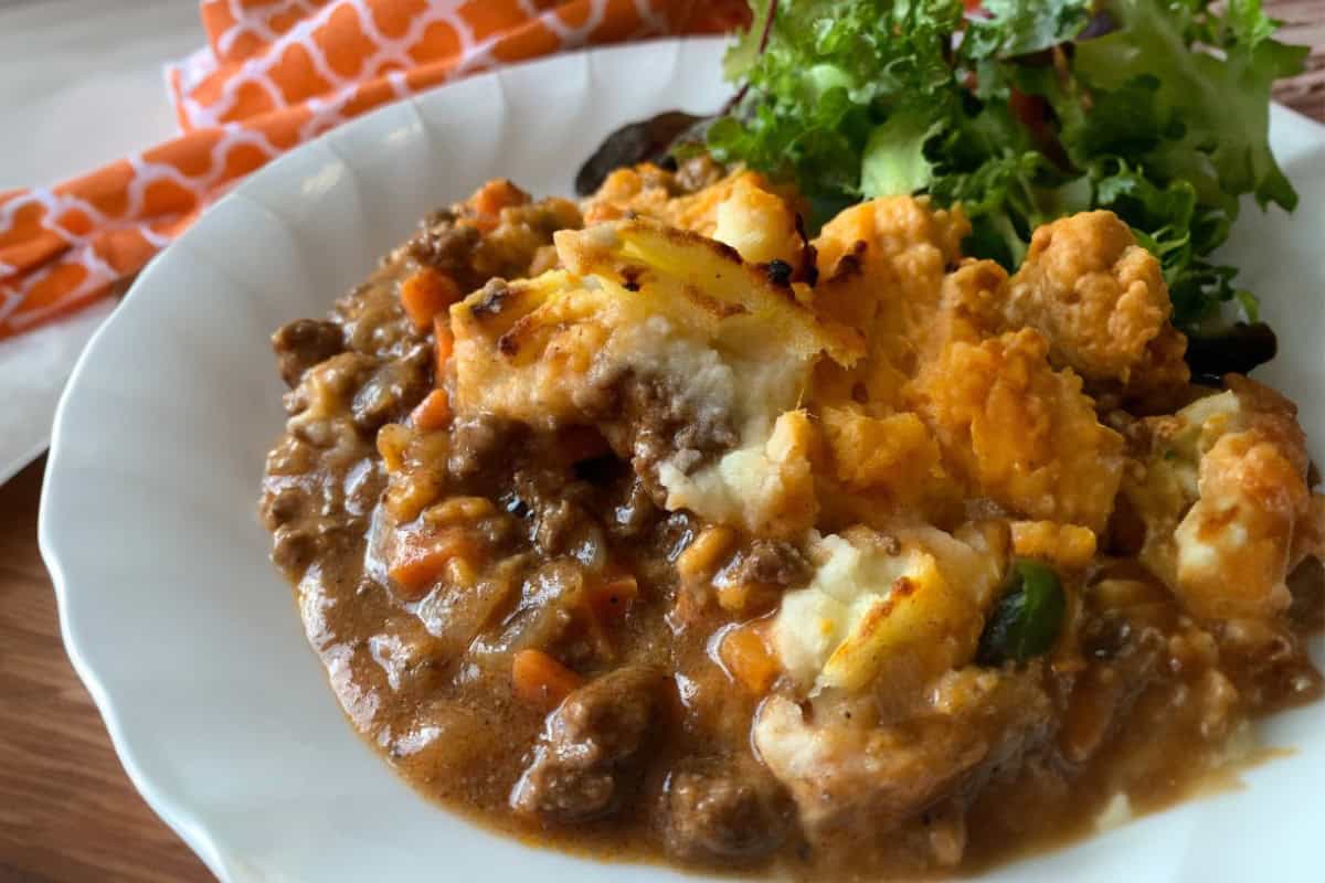 A plate with cottage pie casserole and a side of mixed greens on a white surface, accompanied by an orange-patterned cloth in the background.