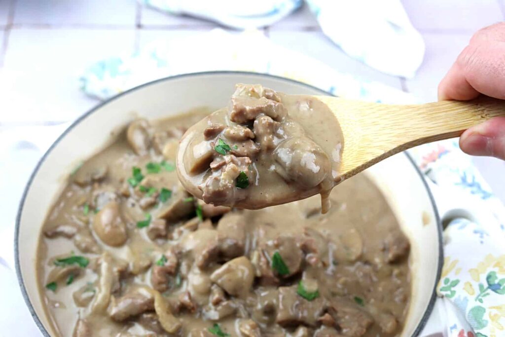 A wooden spoon holds a serving of beef stroganoff with mushrooms and creamy sauce above a pot filled with the dish.