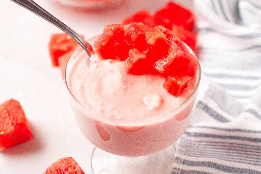 A glass dish filled with pink watermelon mousse, topped with diced watermelon pieces, sits on a white surface next to a striped cloth.