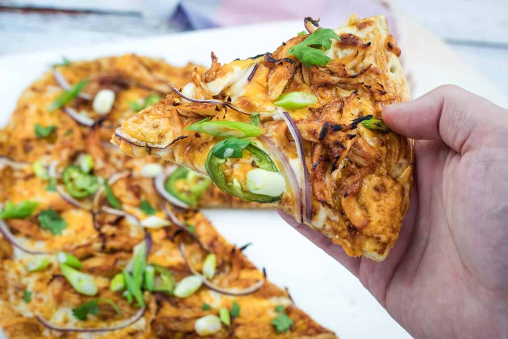 A hand holds a slice of pizza topped with shredded chicken, jalapeño slices, red onions, and green onions; the rest of the pizza is visible on a white surface.
