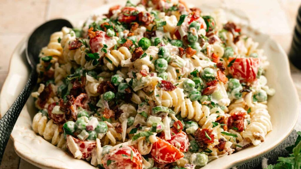 A bowl of rotini pasta salad mixed with peas, cherry tomatoes, bacon bits, chopped herbs, and a creamy dressing, with a black serving spoon beside the dish.