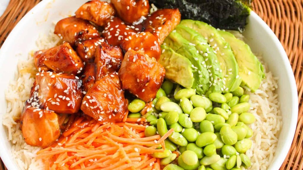 A bowl with rice, glazed chicken pieces, sliced avocado, edamame, shredded carrots, seaweed, and sesame seeds.