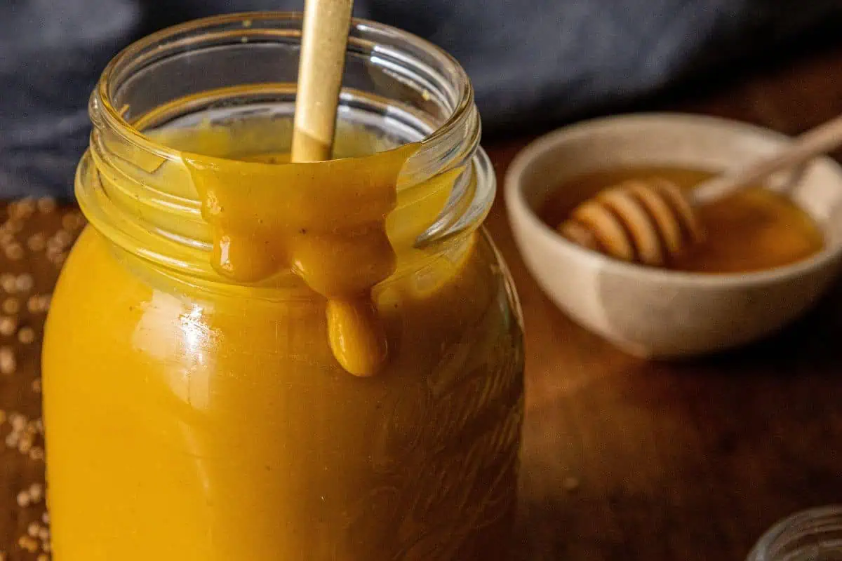 A glass jar filled with thick, golden yellow Carolina Mustard BBQ Sauce, some dripping down the side; a spoon rests inside. In the background, there's a small bowl of honey with a honey dipper—perfect for drizzling over pulled pork.