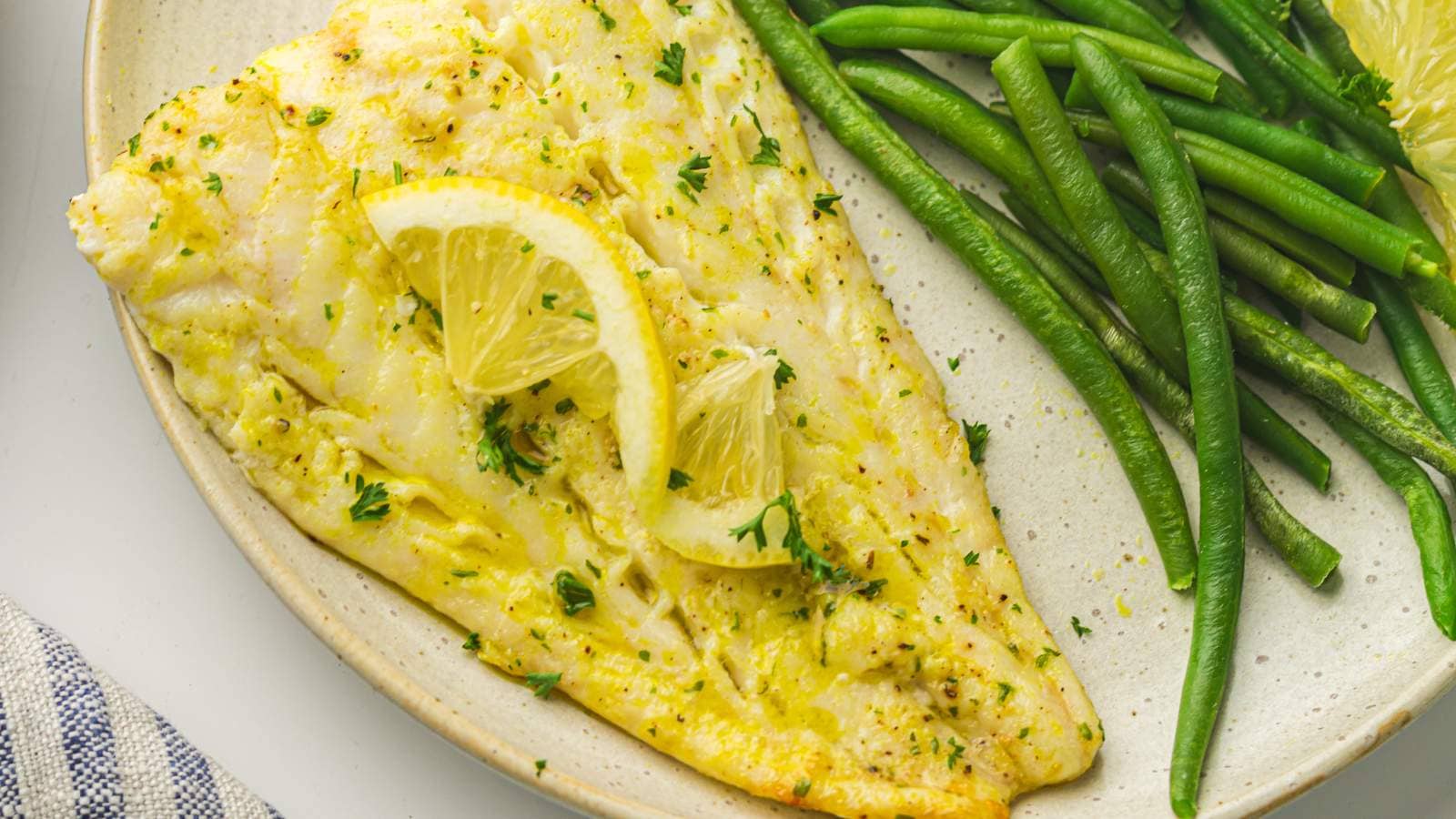 A plate with a cooked white fish fillet garnished with lemon slices and herbs, served alongside green beans.