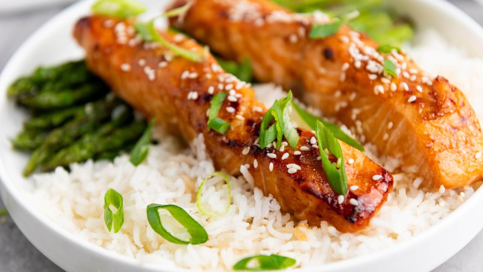 Two glazed salmon fillets topped with sesame seeds and sliced green onions served over white rice with a side of asparagus.