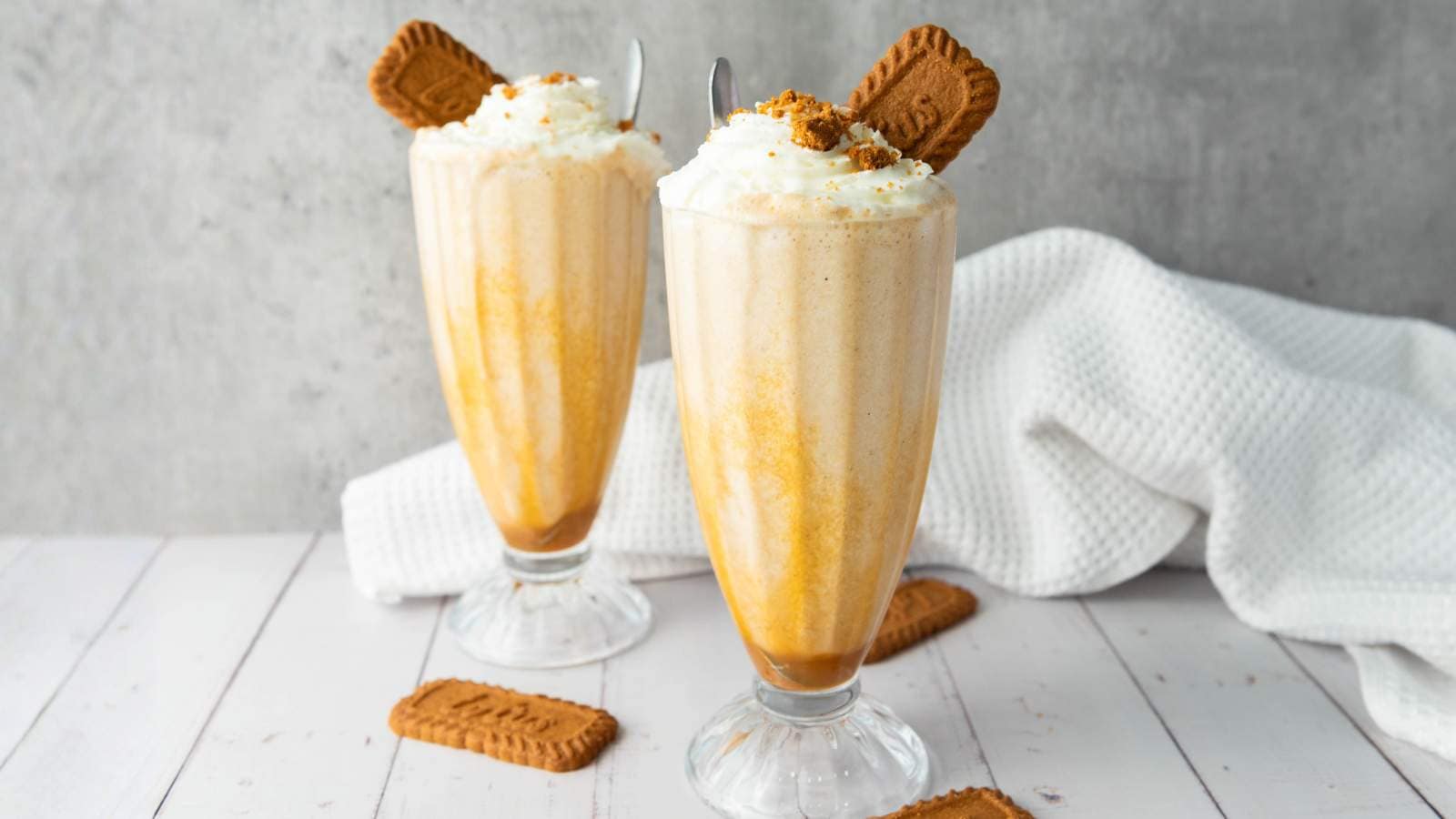 Two tall glasses of creamy Biscoff milkshake topped with whipped cream and biscuits, with extra biscuits on a white wooden surface and a white cloth in the background.