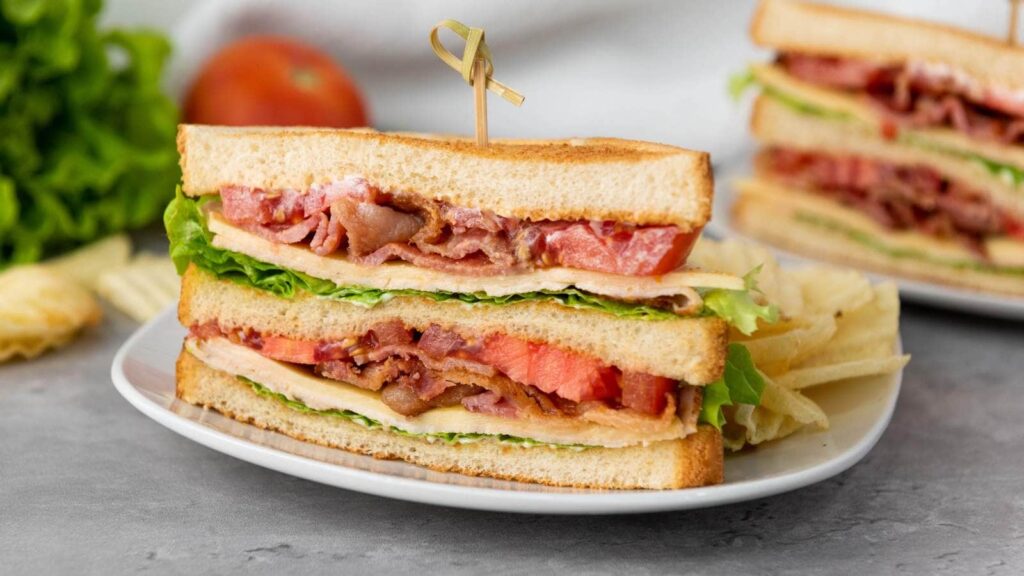 A chicken club sandwich with bacon, lettuce, tomato, and cheese is cut in half and stacked on a plate with potato chips.