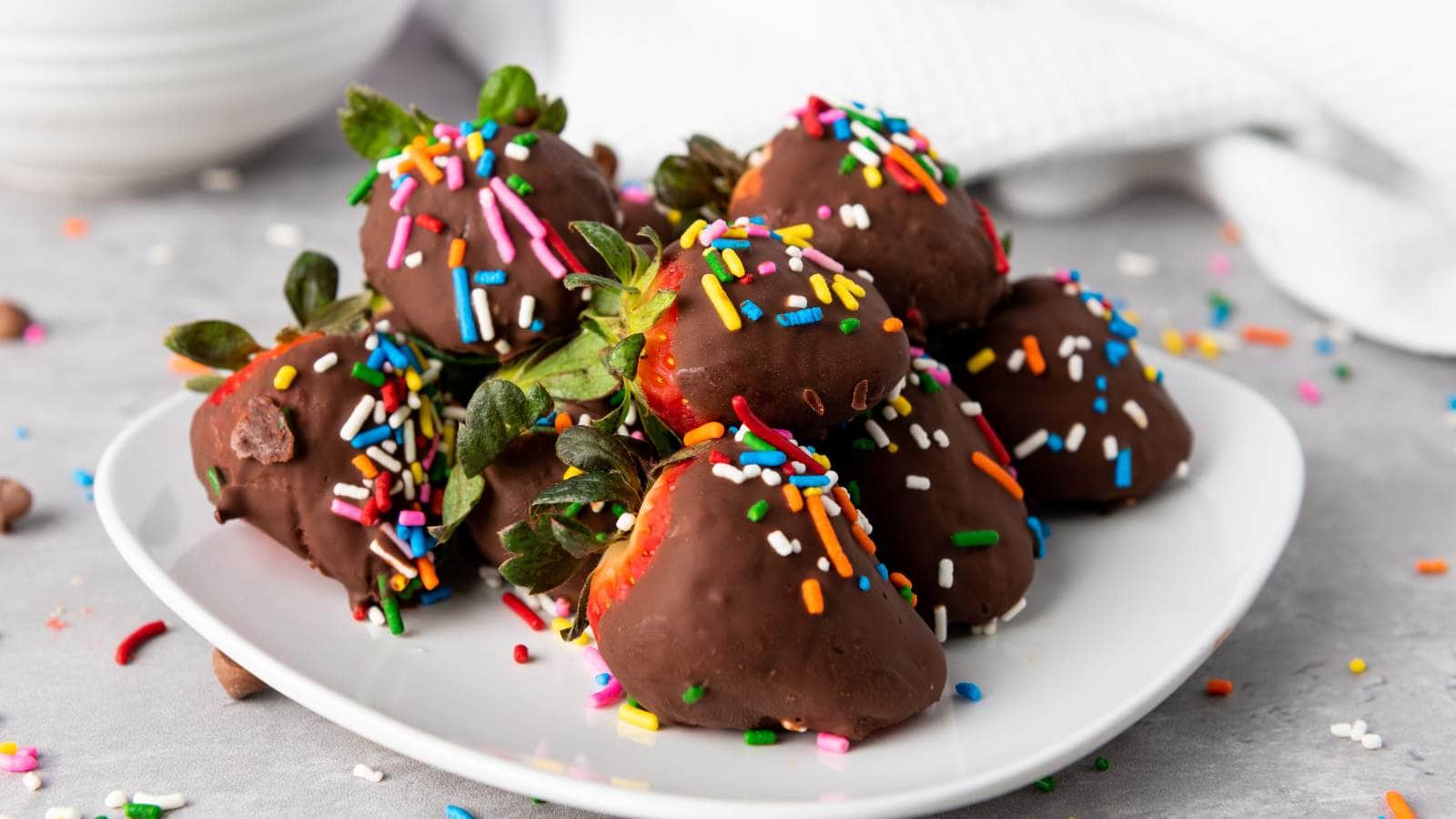 A white plate holds several chocolate-covered strawberries topped with colorful sprinkles, with extra sprinkles scattered on the gray surface nearby.
