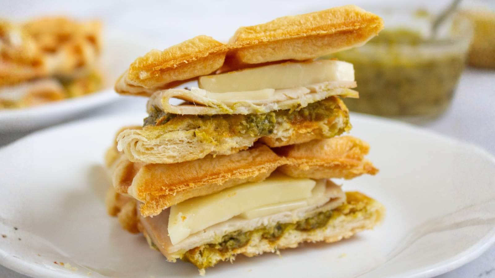 Two croffle waffle sandwiches stacked on a white plate, each filled with slices of cheese, turkey, and a green spread.