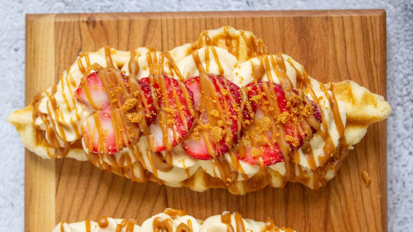 A waffle topped with whipped cream, sliced strawberries, crumbled cookie, and caramel drizzle on a wooden board.