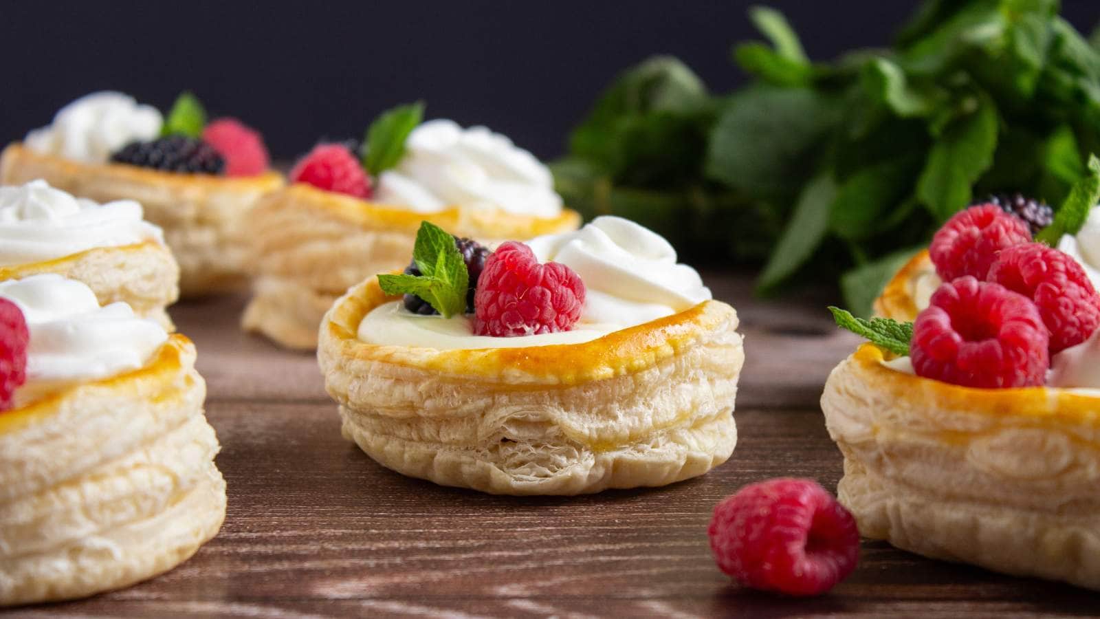Puff pastry shells filled with cream, topped with fresh raspberries, blackberries, and mint leaves, arranged on a wooden surface.