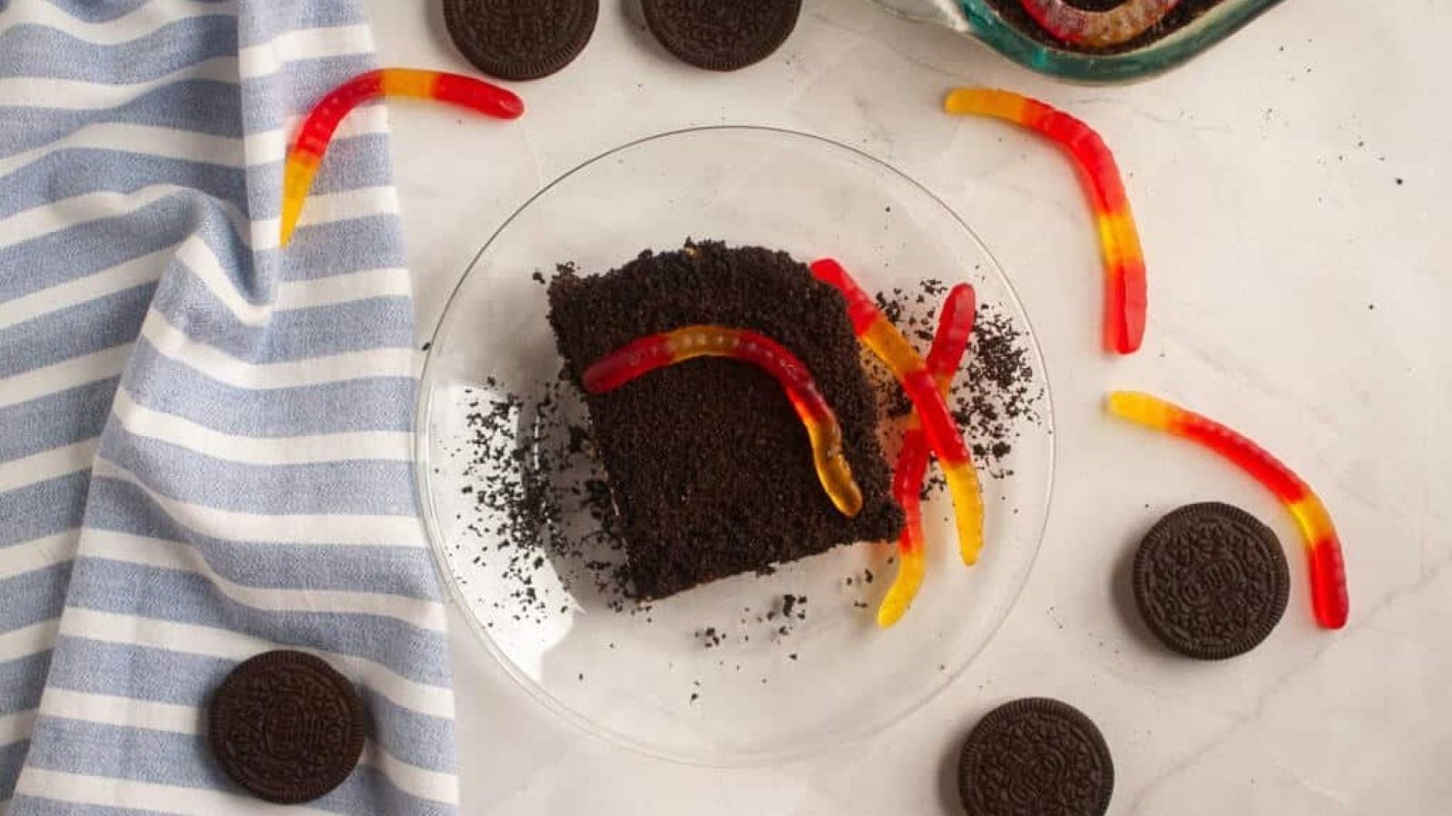 A plate with a slice of dirt cake topped with gummy worms, surrounded by Oreo cookies and a blue-striped cloth on a white surface.