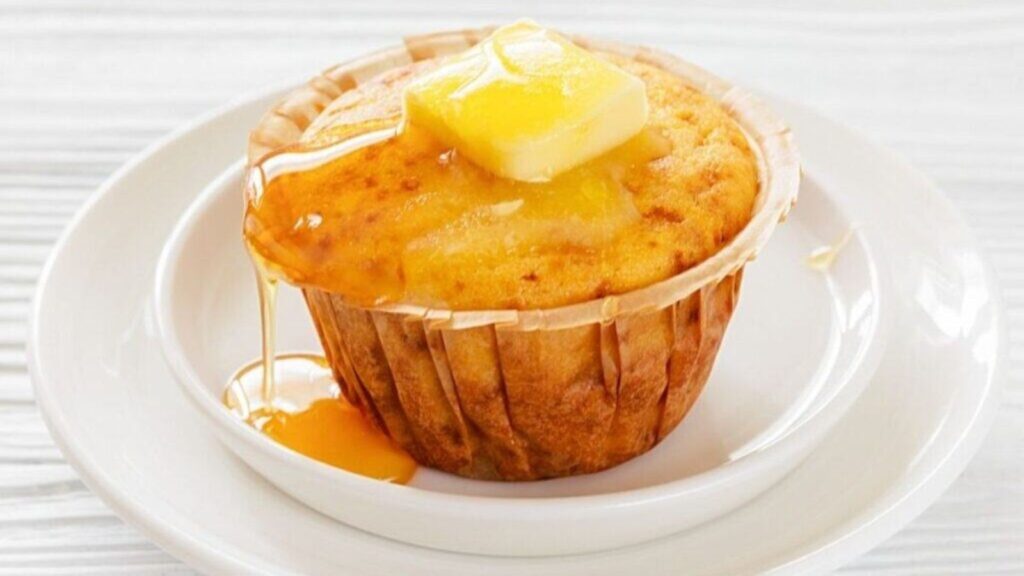 A Honey Cornbread muffin on a white plate, topped with a pat of butter and drizzled with syrup.