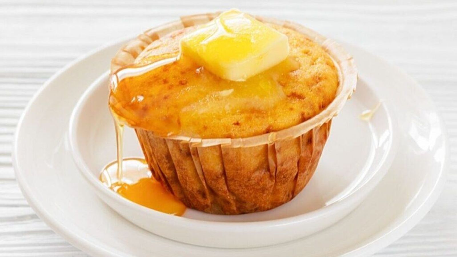 A Honey Cornbread muffin on a white plate, topped with a pat of butter and drizzled with syrup.