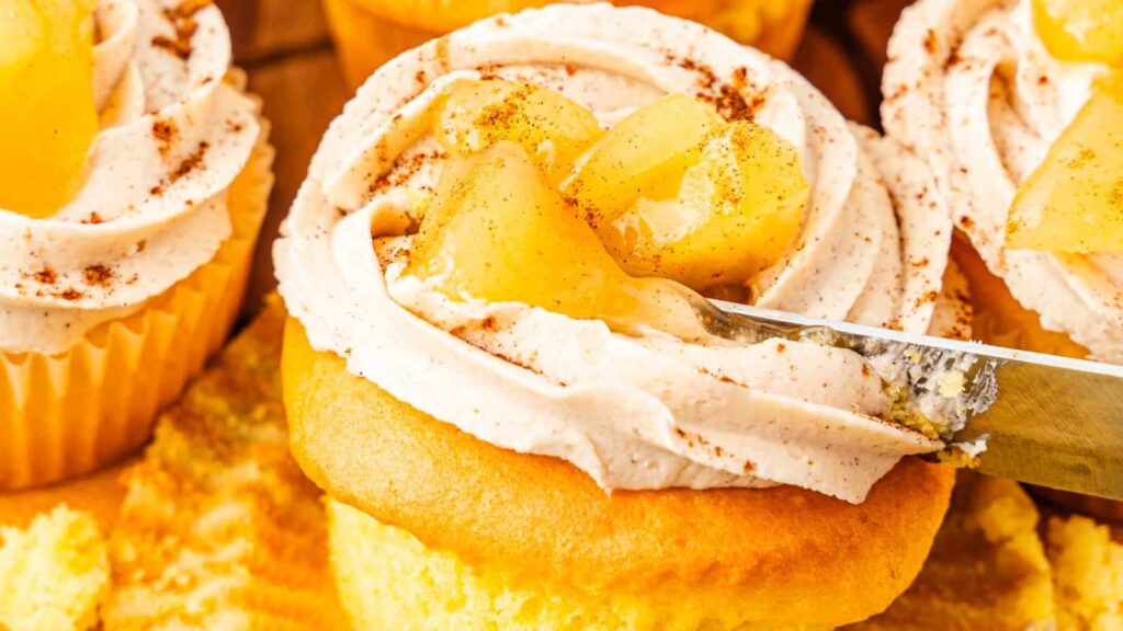 Close-up of cupcakes topped with frosting and apple chunks, sprinkled with cinnamon. A knife is spreading the frosting.