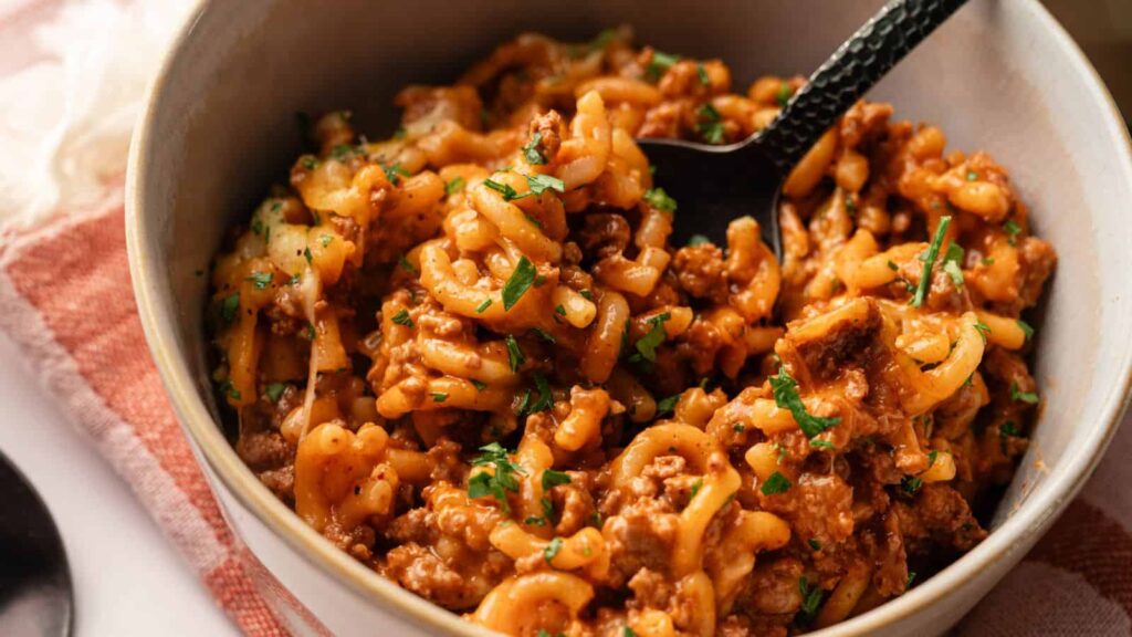 A bowl of macaroni with ground beef in tomato sauce, topped with chopped parsley, with a black spoon in the bowl.