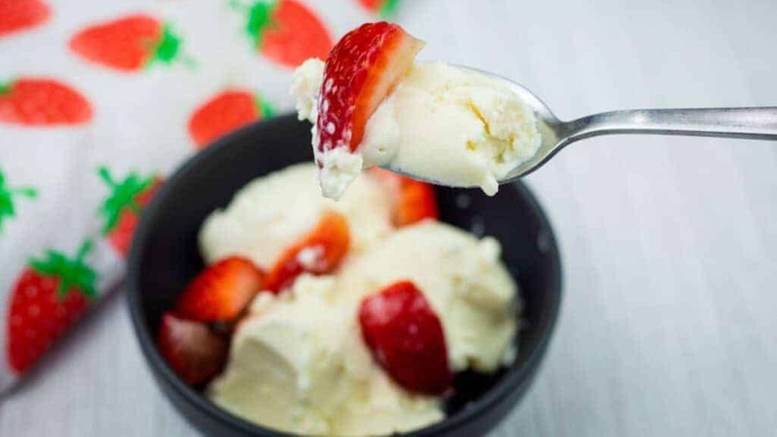 A spoonful of vanilla ice cream and a strawberry held above a bowl with more ice cream and strawberries, next to a strawberry-patterned cloth.