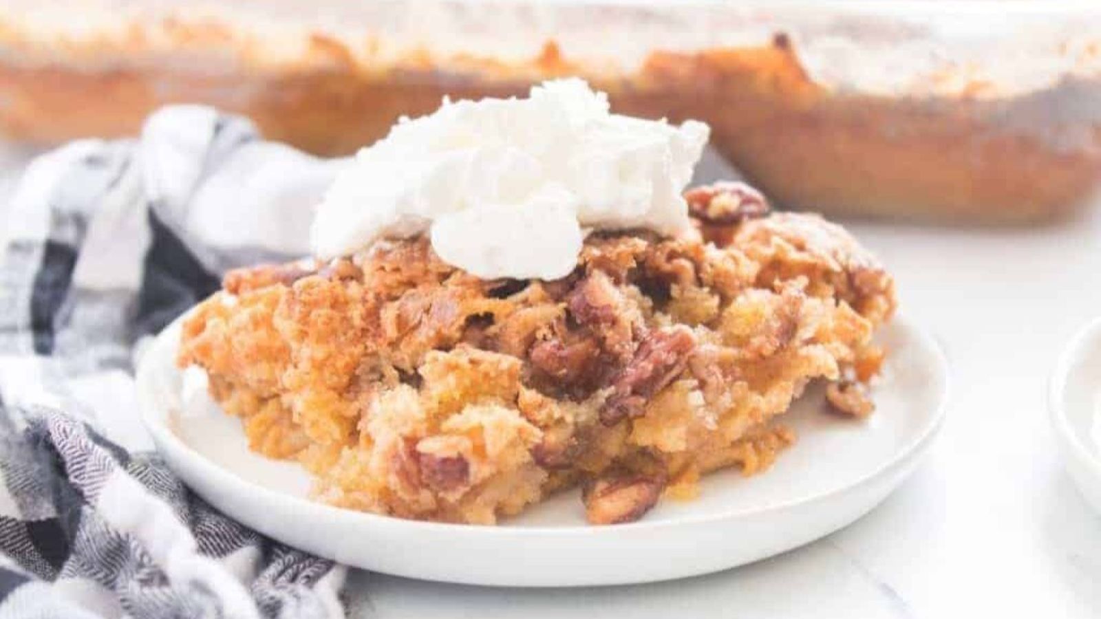 A slice of Pumpkin Crunch Cake served on a white plate with a dollop of whipped cream, napkin beside it, and a baking dish in the background.