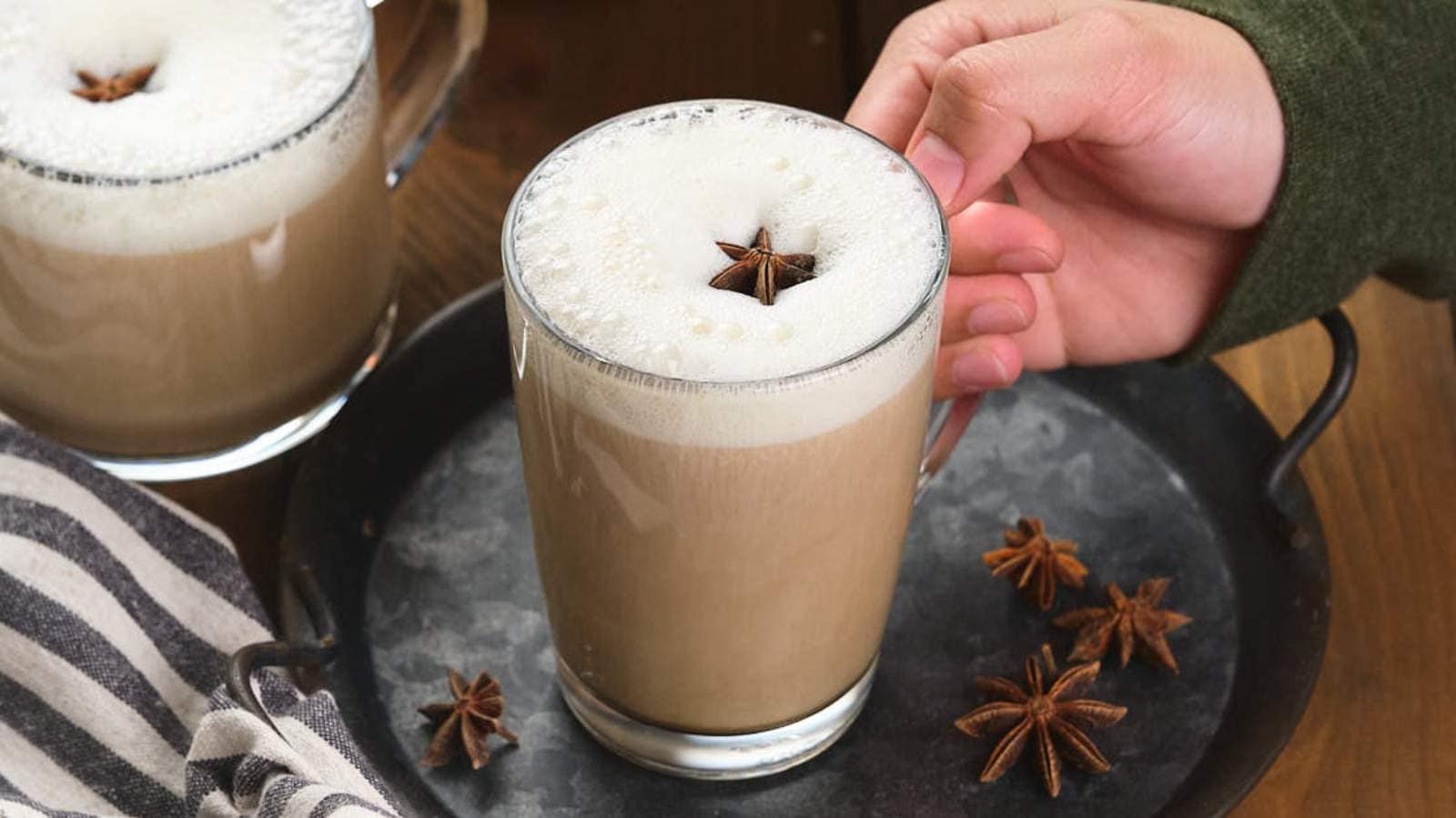 Three glasses of pumpkin chai latte with cinnamon sticks on a clay plate next to a white bowl with brown syrup.
