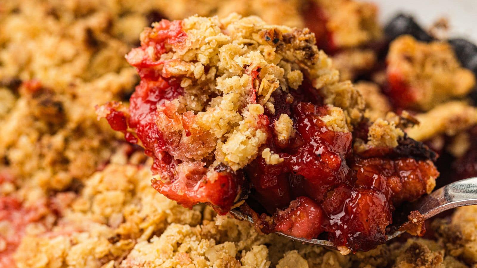 A spoonful of strawberry crumble dessert with a golden oat topping and visible red berry filling.