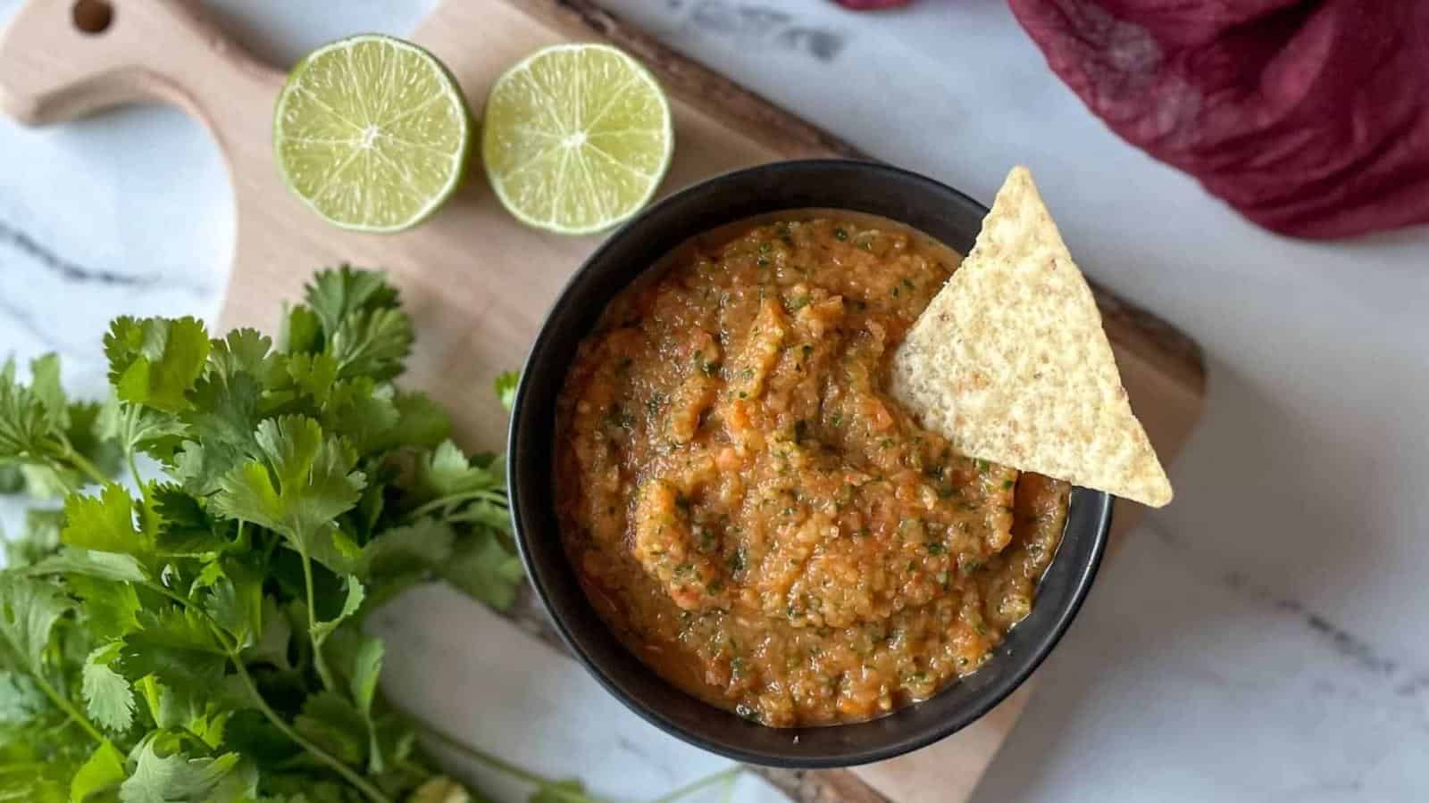 A bowl of pineapple habanero salsa surrounded by cilantro, limes, and a red linen sit on a wooden cutting board.