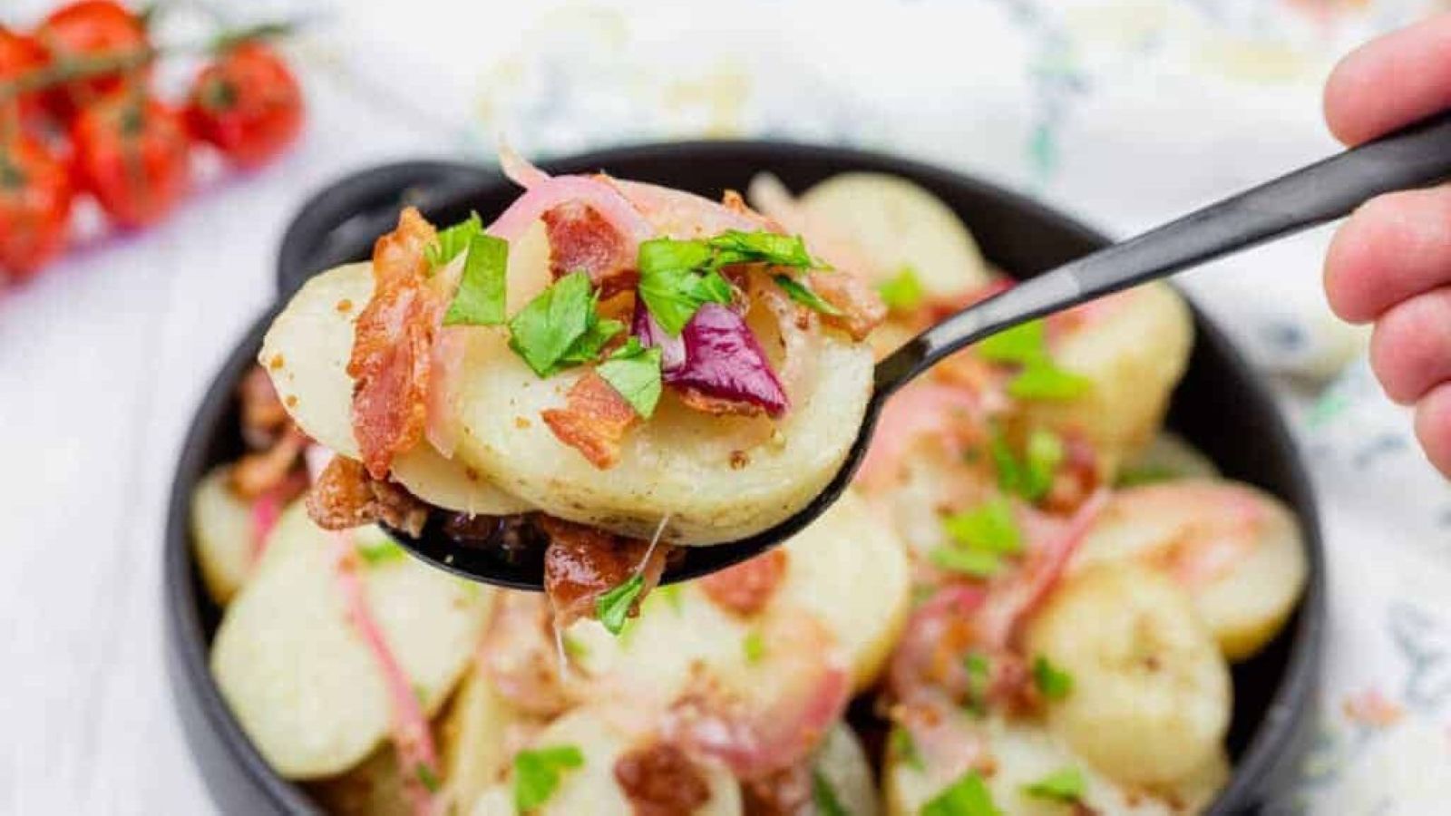 A spoonful of German potato salad topped with bacon, red onion, and chopped parsley held over a bowl of the same salad.