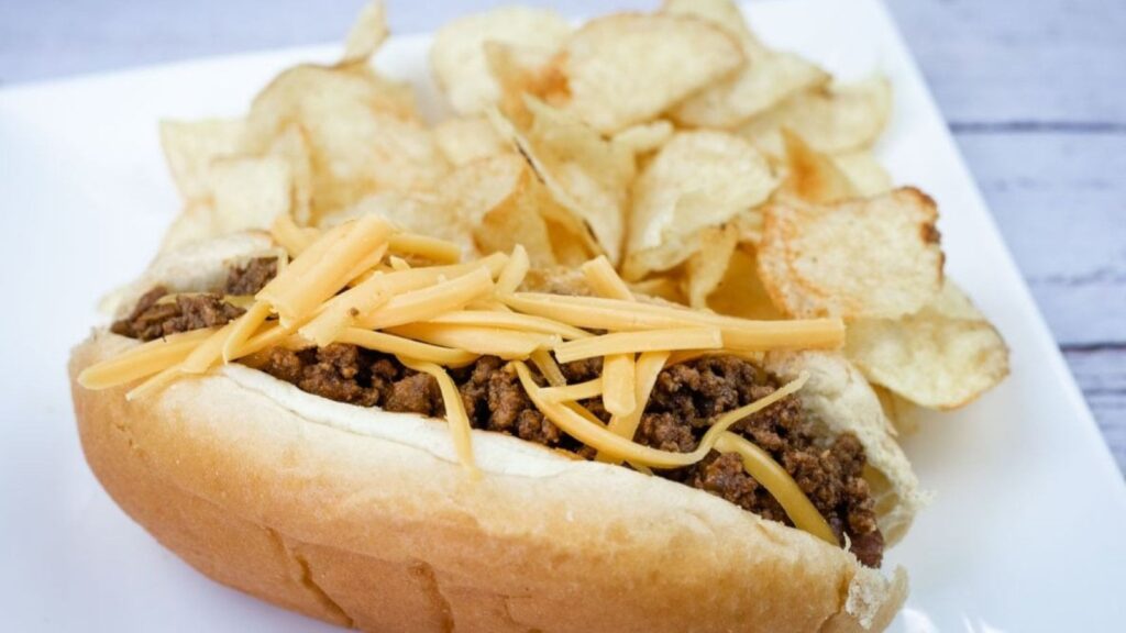 A hot dog chili topped with shredded cheddar, served with potato chips on a white plate.