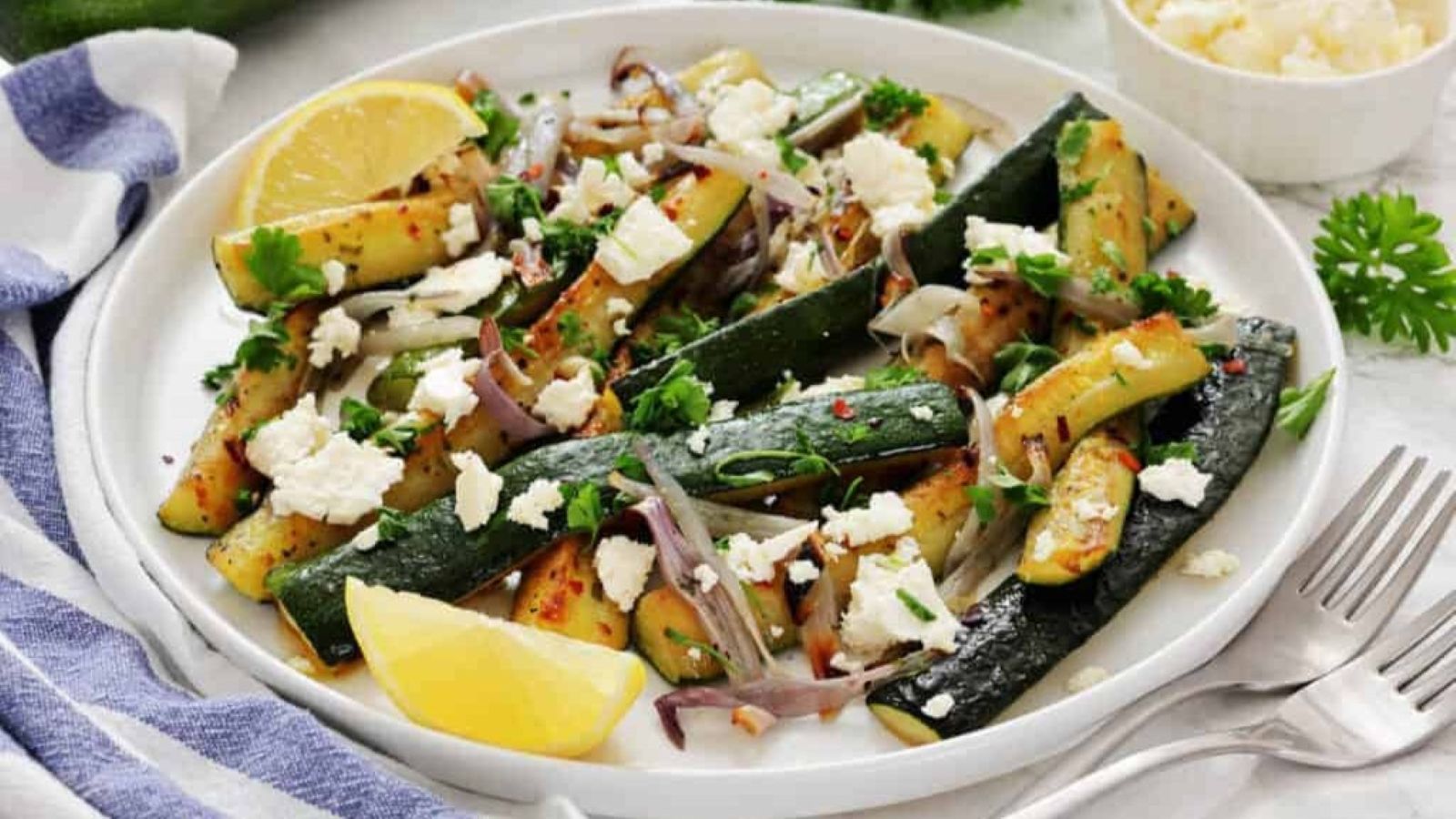 A plate of roasted zucchini and red onions garnished with crumbled cheese, herbs, red pepper flakes, and lemon wedges. A small bowl of cheese and fresh parsley are in the background.