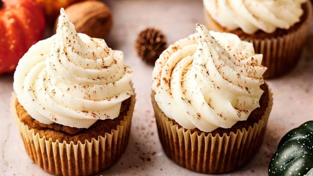 Two cupcakes with white swirled frosting and a sprinkle of cinnamon sit on a light surface, surrounded by fall decorations.