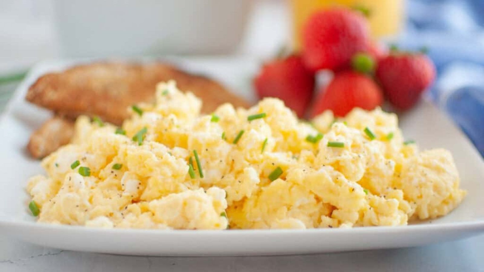A plate of scrambled eggs garnished with chives, served with toast and strawberries, with a glass of orange juice in the background.