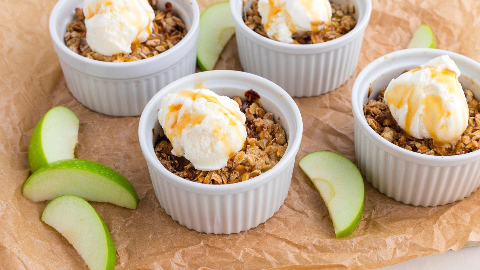 Four ramekins filled with apple crisp topped with ice cream and caramel sauce, placed on brown parchment paper with green apple slices.