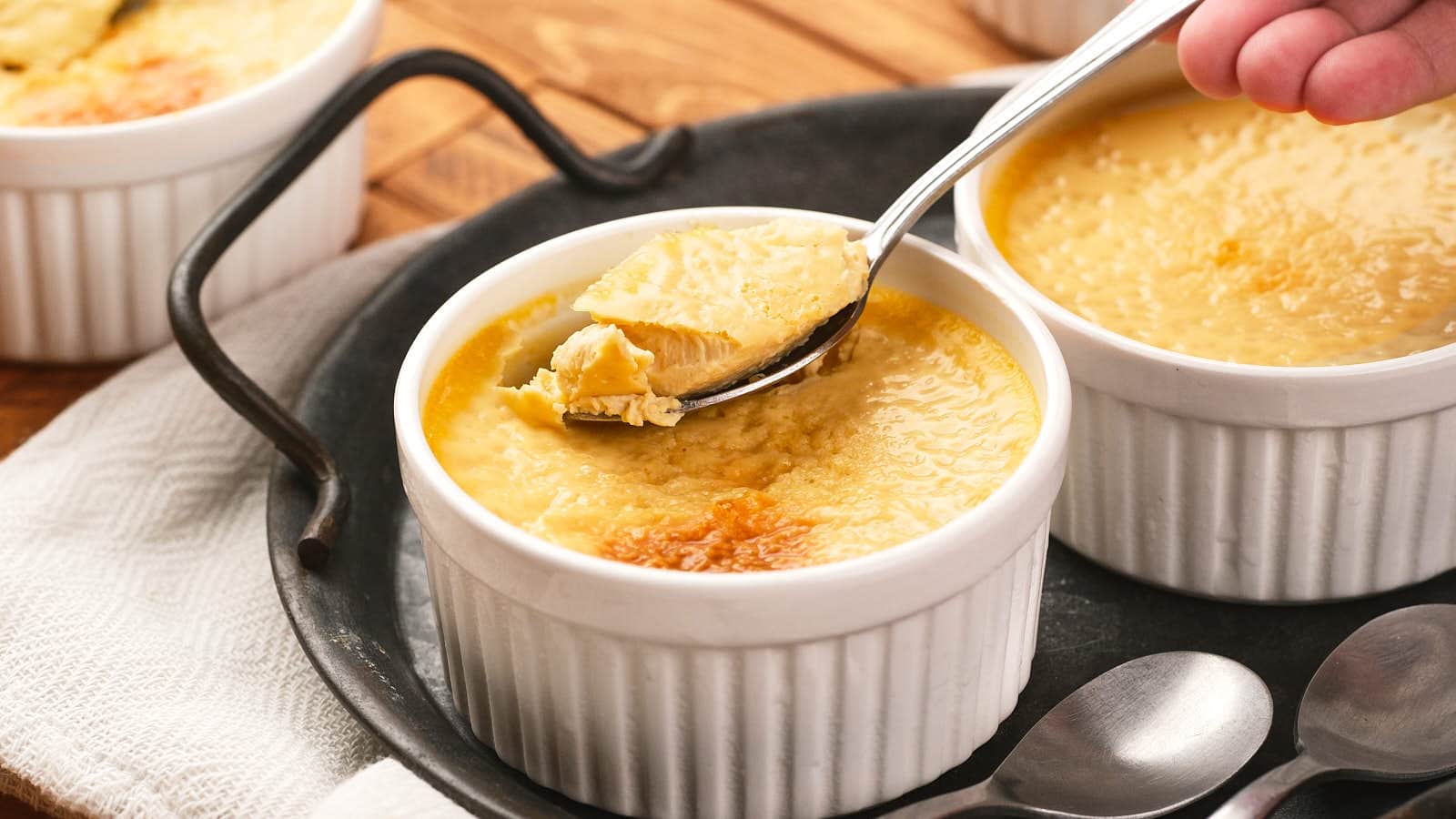 A hand holds a spoon scooping baked custard from a white ramekin, with two more ramekins and spoons on a tray.